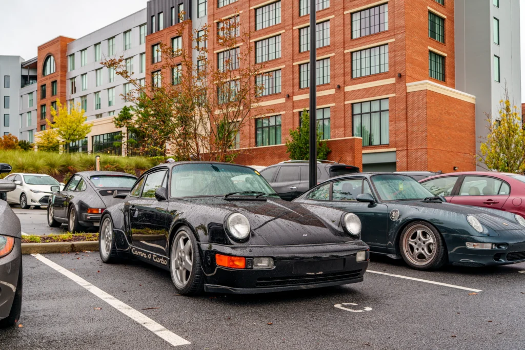Black, green, and gray Porsche 911 coupes sit in a wet parking lot outside a multistory brick building.