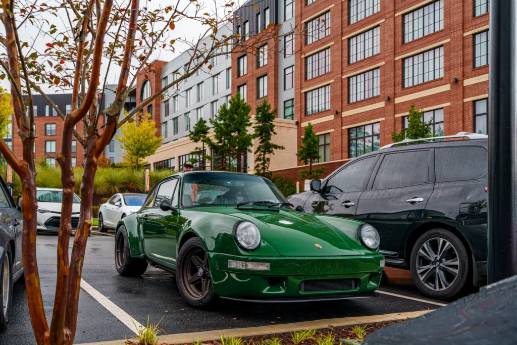 Green Porsche 911 parked next to a black SUV in an urban parking lot with a brick building in the background