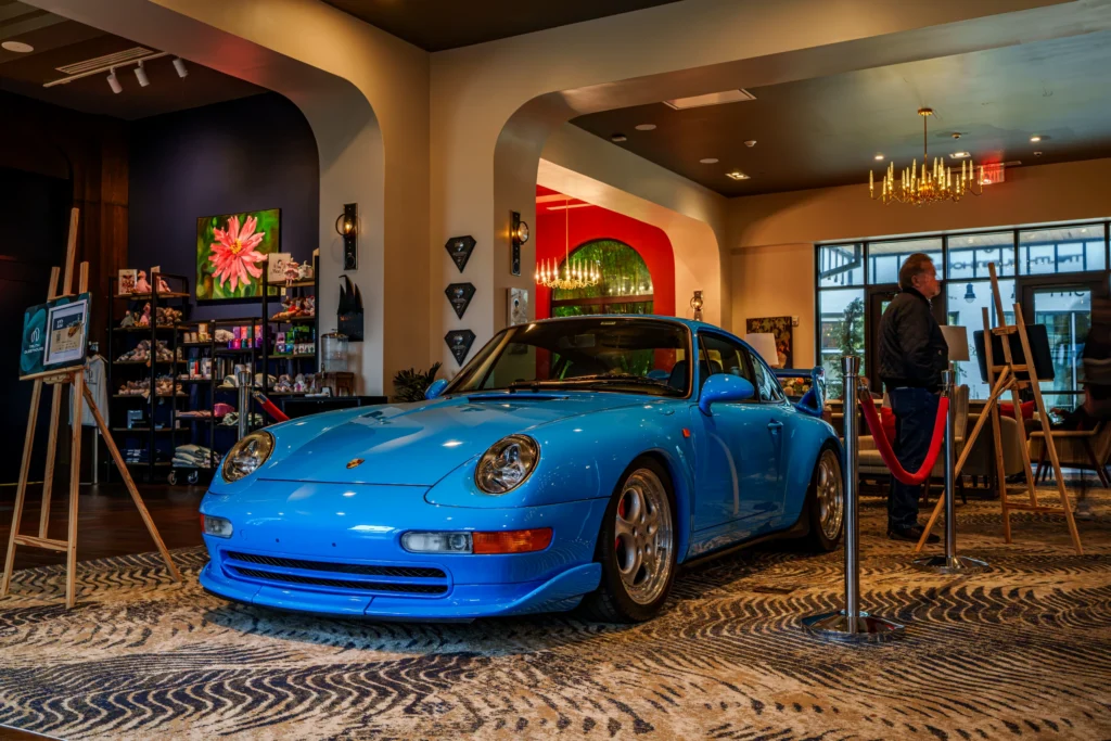 Blue Porsche 911 displayed indoors with a man standing nearby and room decor visible