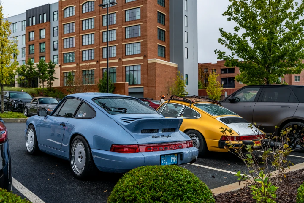 Blue Porsche 911 and yellow Porsche 911 parked side by side in an urban parking lot with brick buildings and greenery
