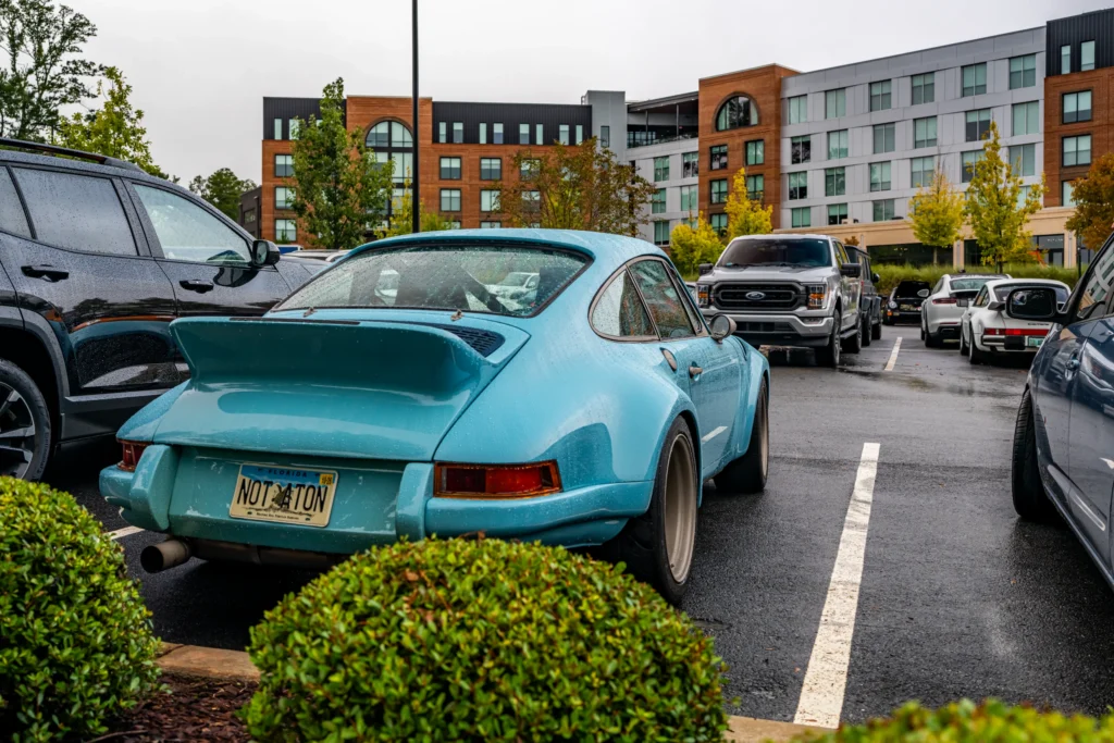 Light blue Porsche 911 parked in a lot on a rainy day with other vehicles including a gray Ford pickup and silver Porsche