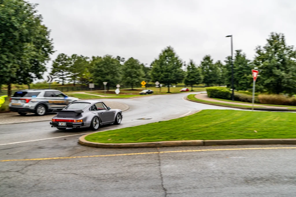 Silver Porsche 911 driving on a curved road with a sheriff SUV nearby and trees in the background