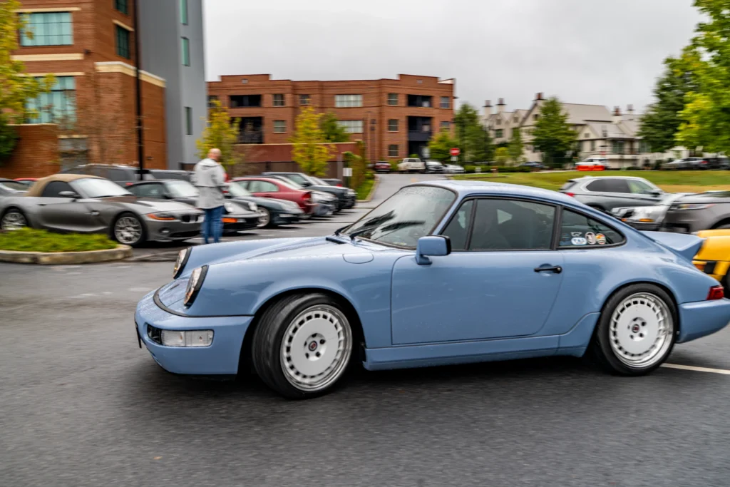 Light blue Porsche 911 with rear spoiler in parking lot on cloudy day with other cars and person nearby