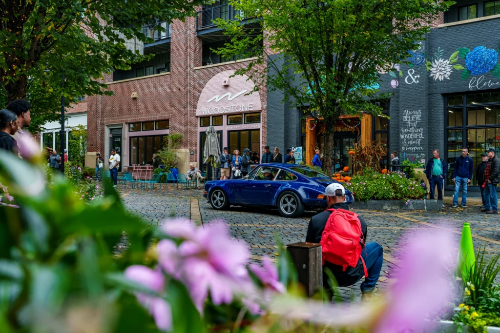 Blue Porsche 911 parked on cobblestone street with people standing near a cafe and colorful flowers in foreground