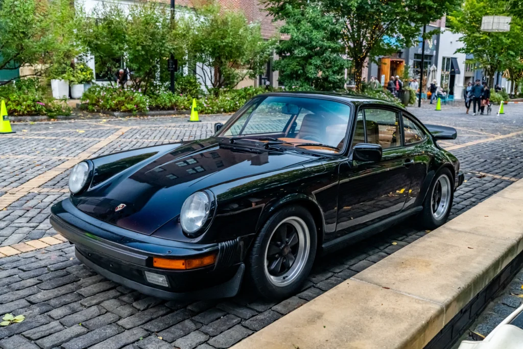 Black Porsche 911 parked on a cobblestone street in an urban area with trees and pedestrians in the background