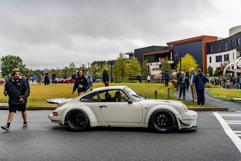 A white Porsche 911 with widened fenders and black wheels sits beside attendees at an outdoor car event.