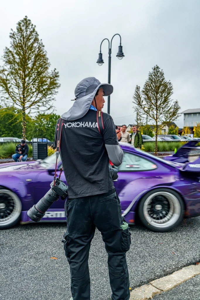 A photographer carrying cameras stands beside a purple Porsche 911 at an outdoor car gathering.