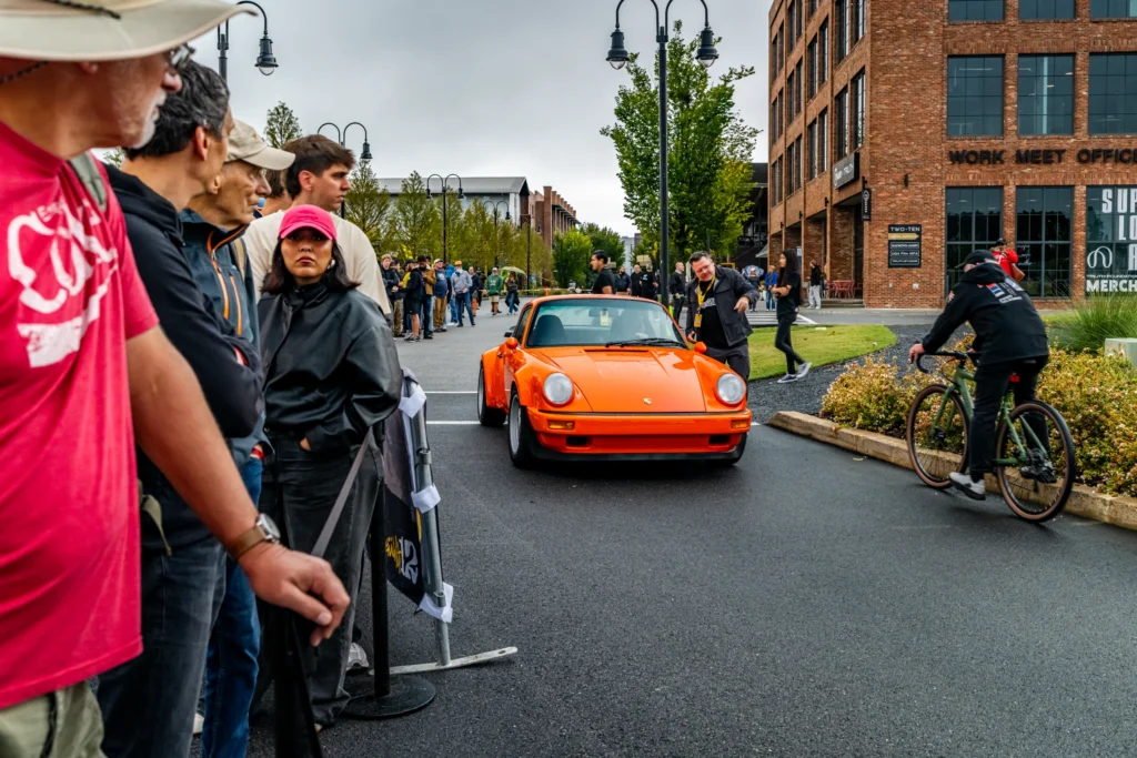 Orange Porsche 911 parked on a street with people lined up along the sidewalk and a cyclist riding past on the right.