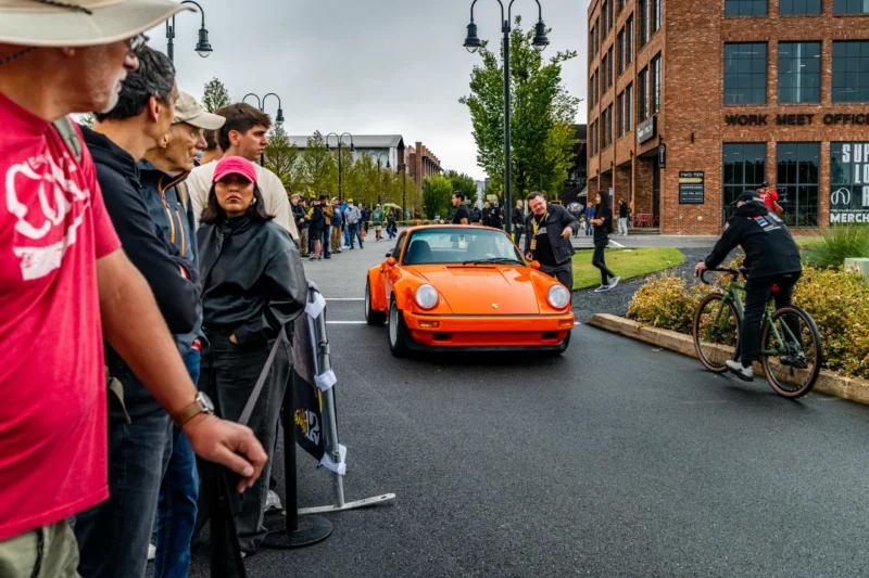 Orange Porsche 911 parked on a street with people lined up along the sidewalk and a cyclist riding past on the right.