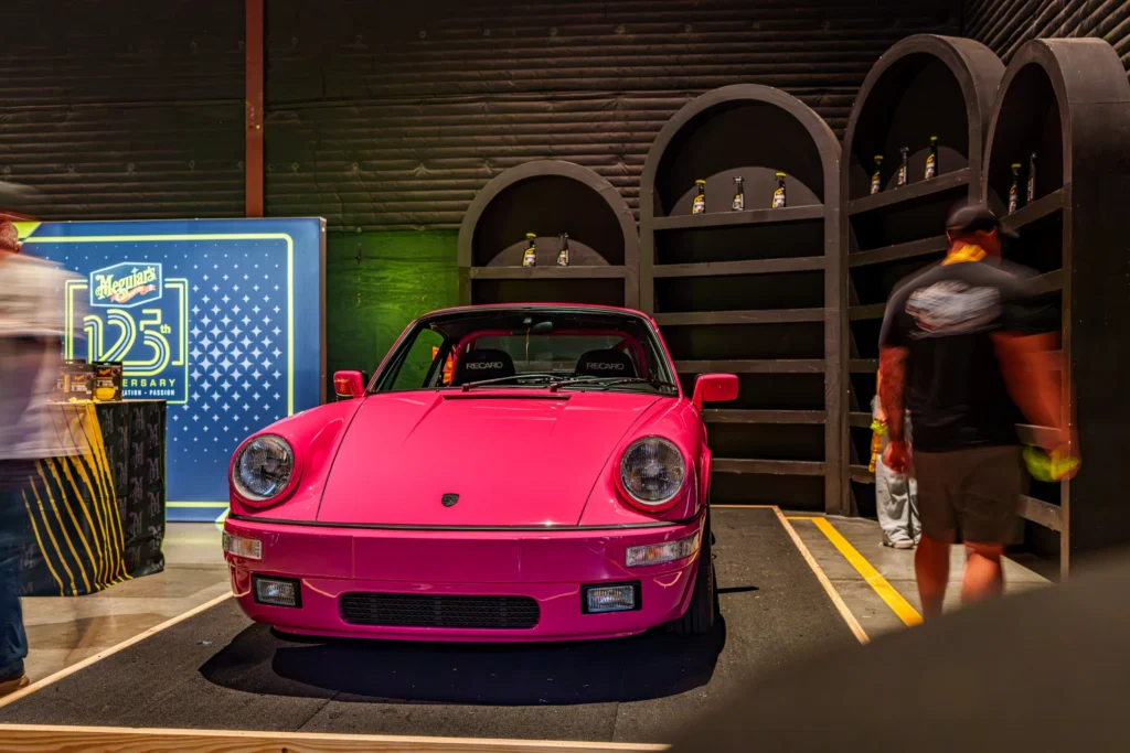 Front view of a pink Porsche 911 parked indoors near display shelves and signs with blurred people nearby