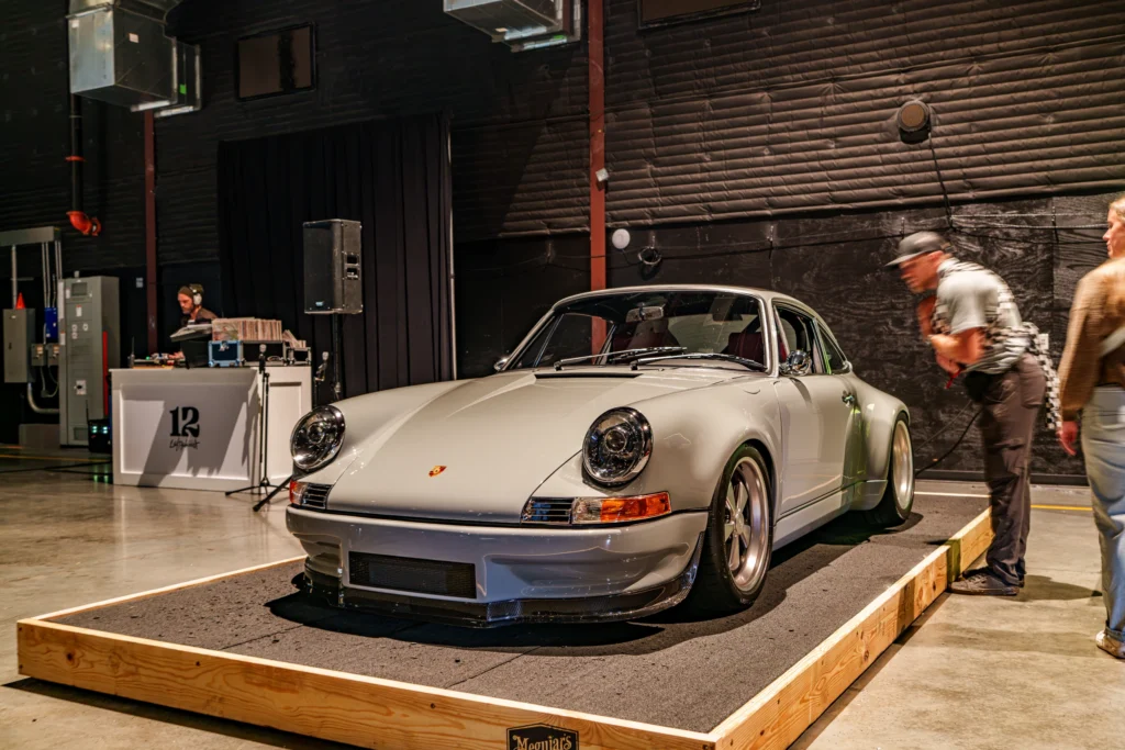 White Porsche 911 displayed indoors on a raised platform with people observing near it and a DJ booth in the background.