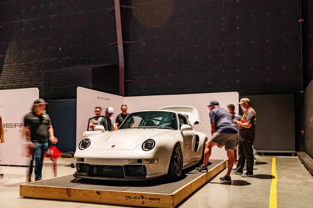 White Porsche 911 on raised platform indoors with several people standing around it and black background walls