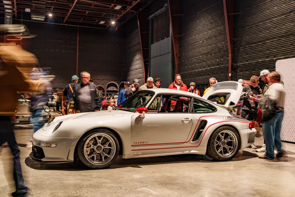 White Porsche 911 with red side mirror and accents indoors surrounded by people at car event