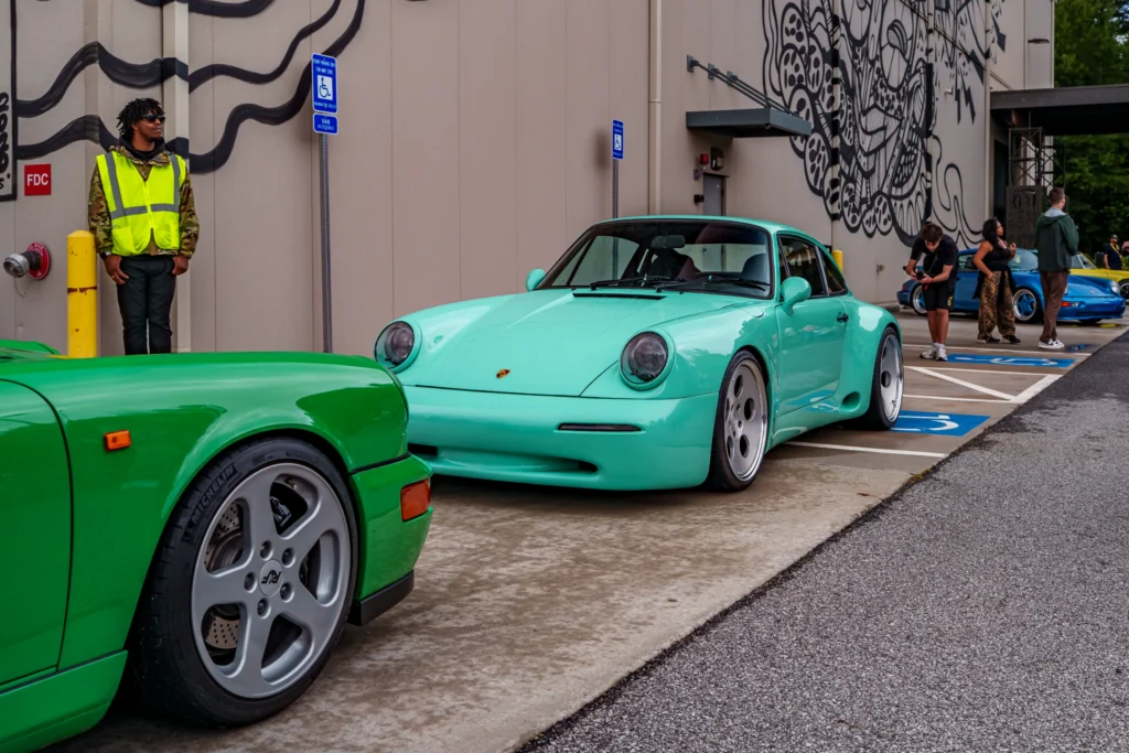 Mint green Porsche 911 and bright green Porsche 964 parked in an outdoor lot near a wall with black mural art