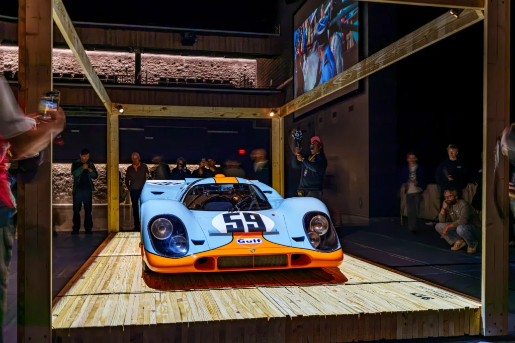 Light blue and orange Porsche race car with number 59 on display inside a dimly lit room with wooden beams and spectators