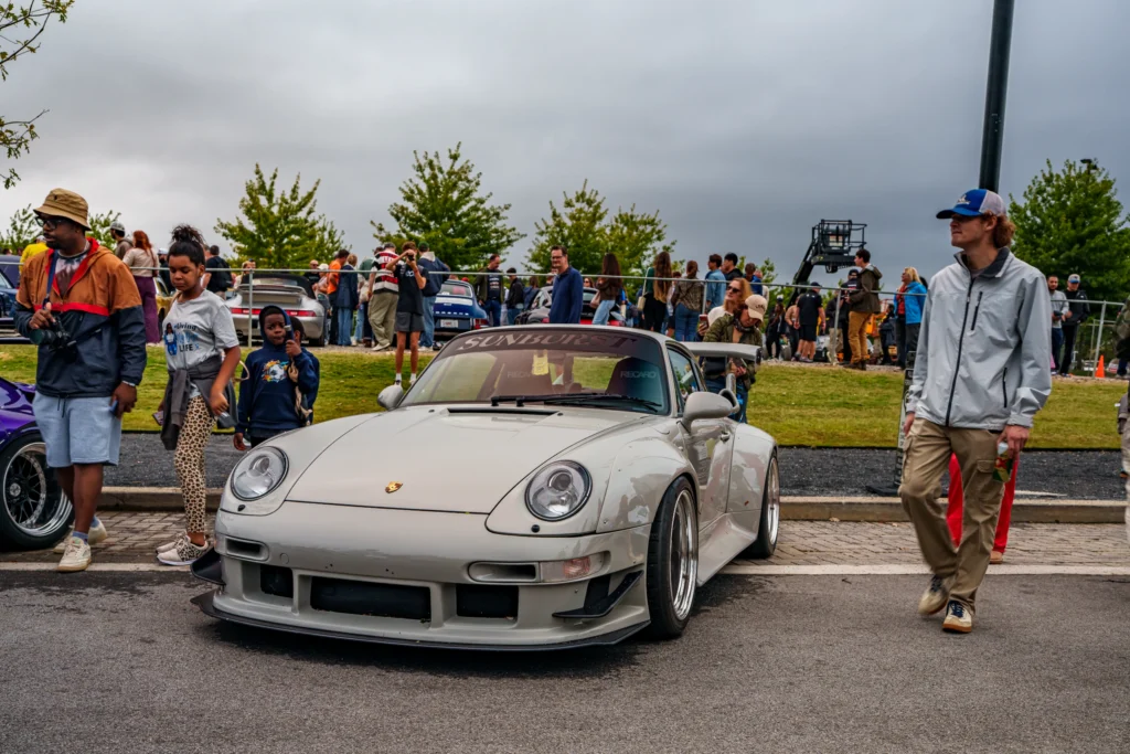 White Porsche 911 with rear wing parked among people at an outdoor car event under cloudy skies.