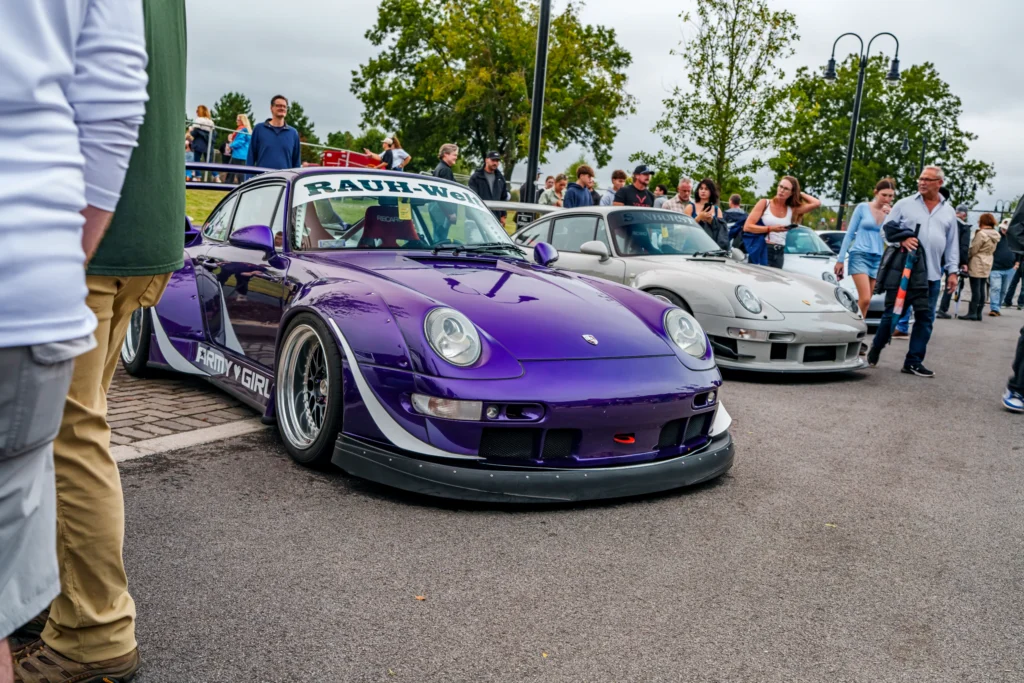 Purple Porsche 911 with Rauh-Welt BEGRIFF decals parked beside a light gray Porsche 911 with Sunburst decal and crowd in