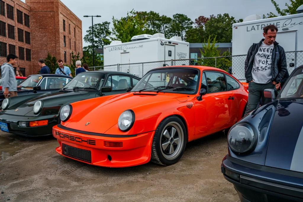 Three Porsche 911 cars in green, orange, and dark blue parked side by side at an outdoor car event with people in the