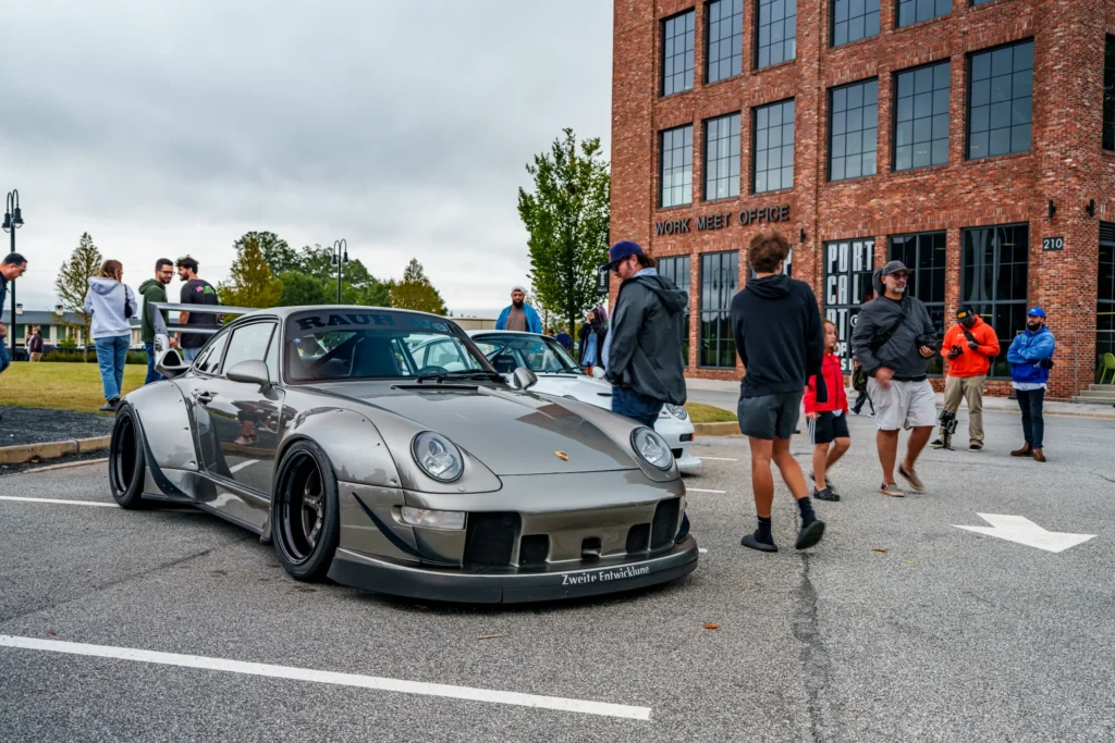 Gray Porsche 911 with RAUH-Welt lettering on windshield parked at car meet with people and brick building in background
