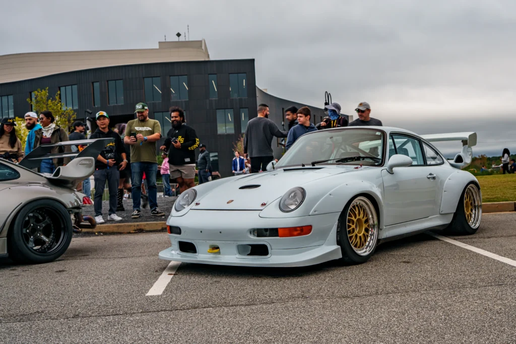 White Porsche 911 with wide body and large rear wing parked at a car event with people in background under overcast sky.