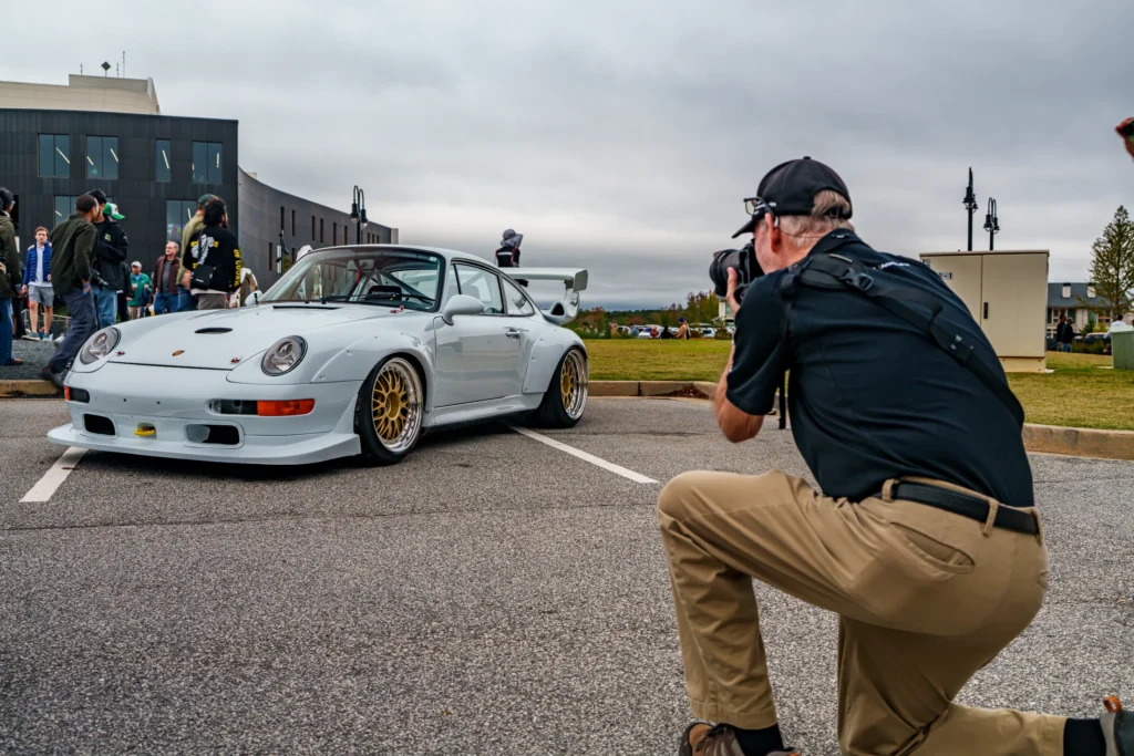 White Porsche 911 parked in a lot with a man photographing it, people and a modern building are in the background under