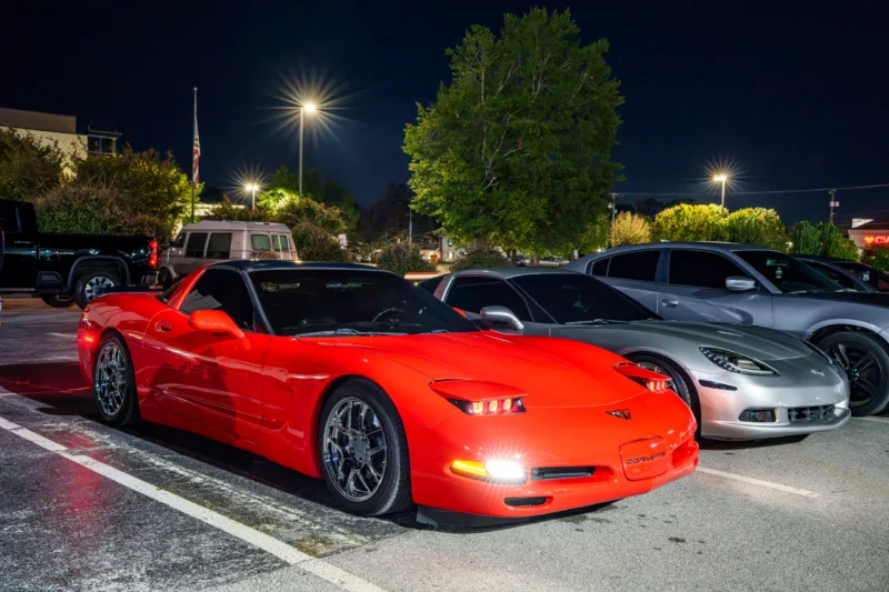 Two Chevrolet Corvettes, one red and one silver, parked side by side at night in an outdoor lot under streetlights.