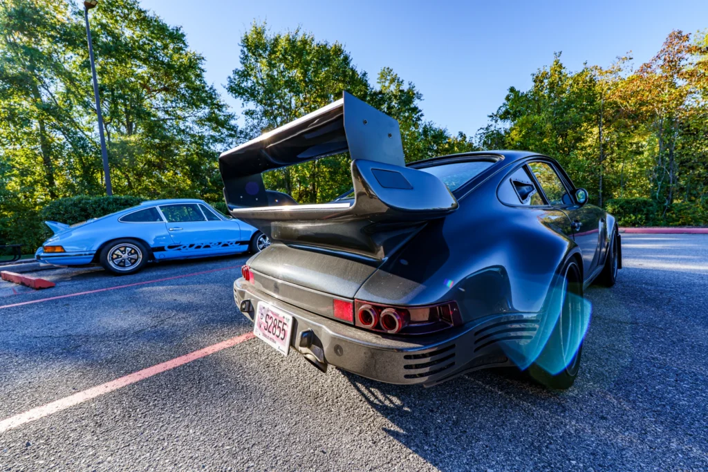 Black Porsche 911 with large rear wing in parking lot with blue Porsche 911 Carrera behind it under clear sky