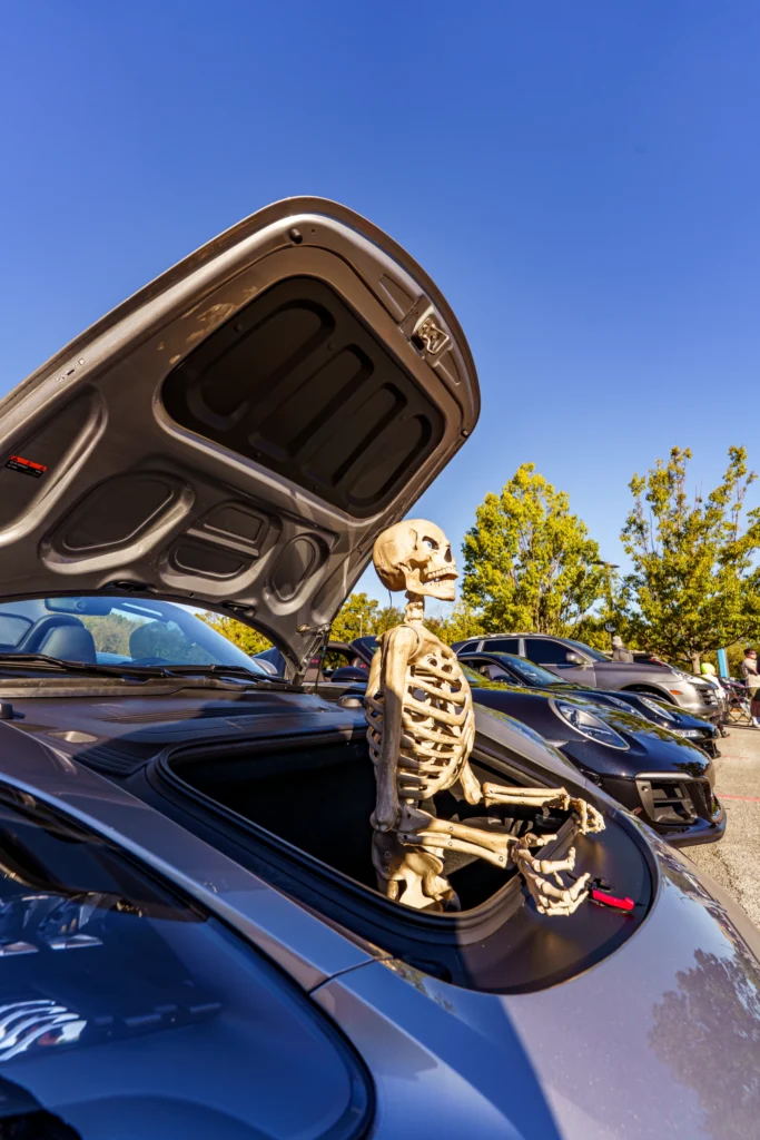 Skeleton sitting in the front trunk of a silver Porsche with other cars and trees in a parking lot under blue sky