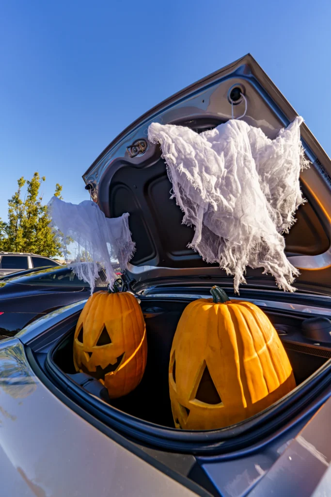 Open trunk of a silver car with two jack-o'-lanterns and white fabric hanging from the trunk lid, under clear blue sky