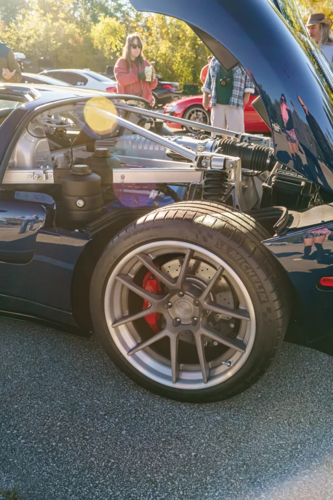Open rear engine bay showing suspension and tire of a dark blue Ford GT at an outdoor car event with people in the background