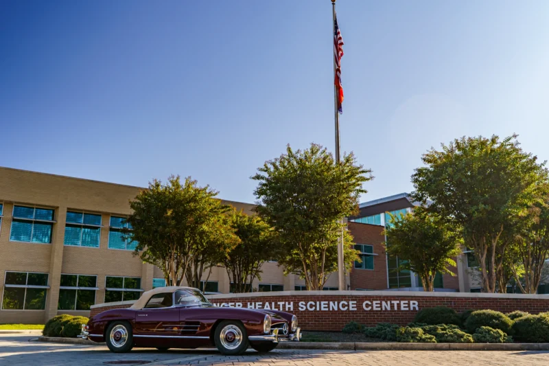 Dark red Mercedes-Benz 300SL convertible parked in front of a health science center building with trees and a flagpole