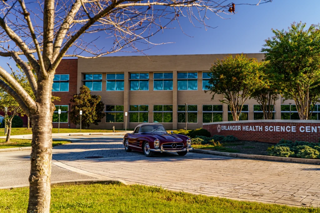 Maroon Mercedes-Benz 300SL convertible parked on pavement outside a medical science building on a sunny day