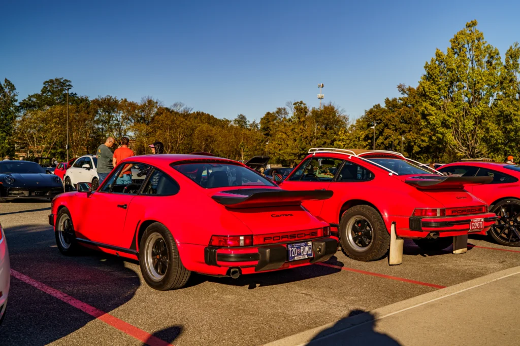 Two red Porsche 911 Carrera cars parked side by side at an outdoor car event with trees in the background under clear blue