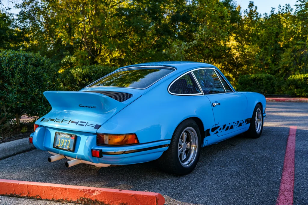 Blue 1973 Porsche 911 Carrera RS parked with rear and driver's side visible, surrounded by green trees in daylight