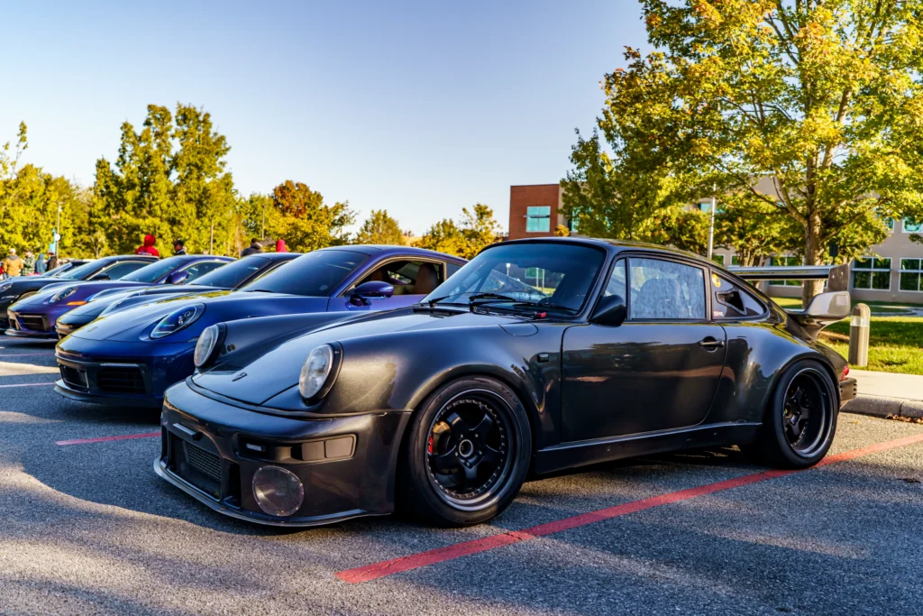 Several Porsche 911 models lined up in a row at an outdoor car event with trees and a building in the background.