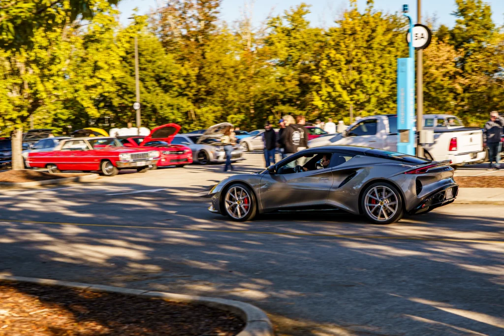 A gray Lotus Emira drives past parked show cars and attendees in a tree-lined parking lot.