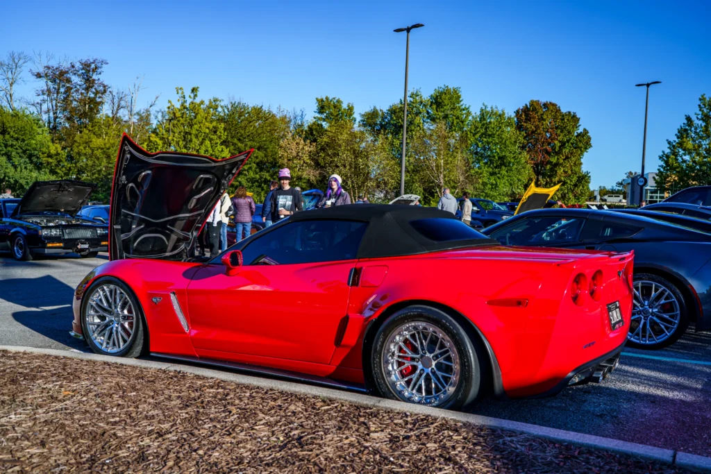 Red Chevrolet Corvette convertible parked at a daytime car event with hood open and people in the background