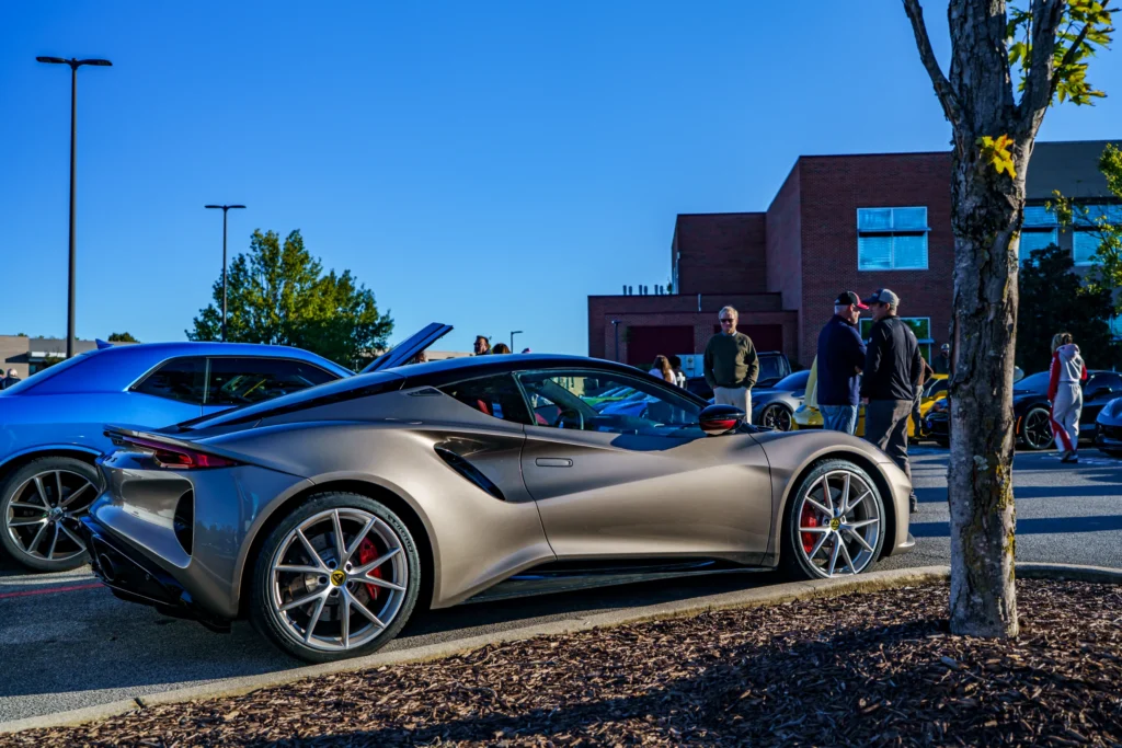 Bronze Lotus Emira coupe parked in a car meet lot with a blue Dodge Challenger behind and attendees in the background