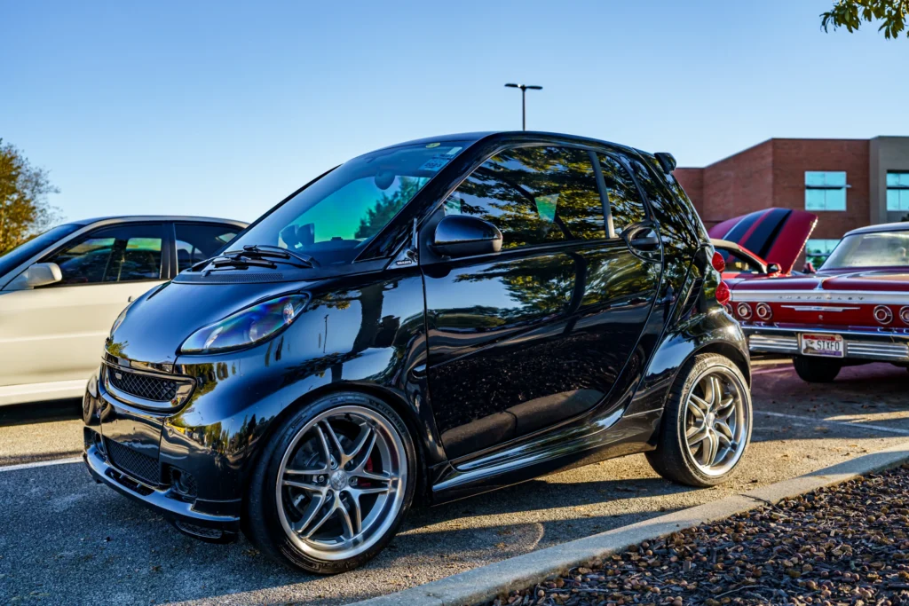 Black Smart car parked in a lot with a white sedan and a red Pontiac Bonneville with raised hood in the background