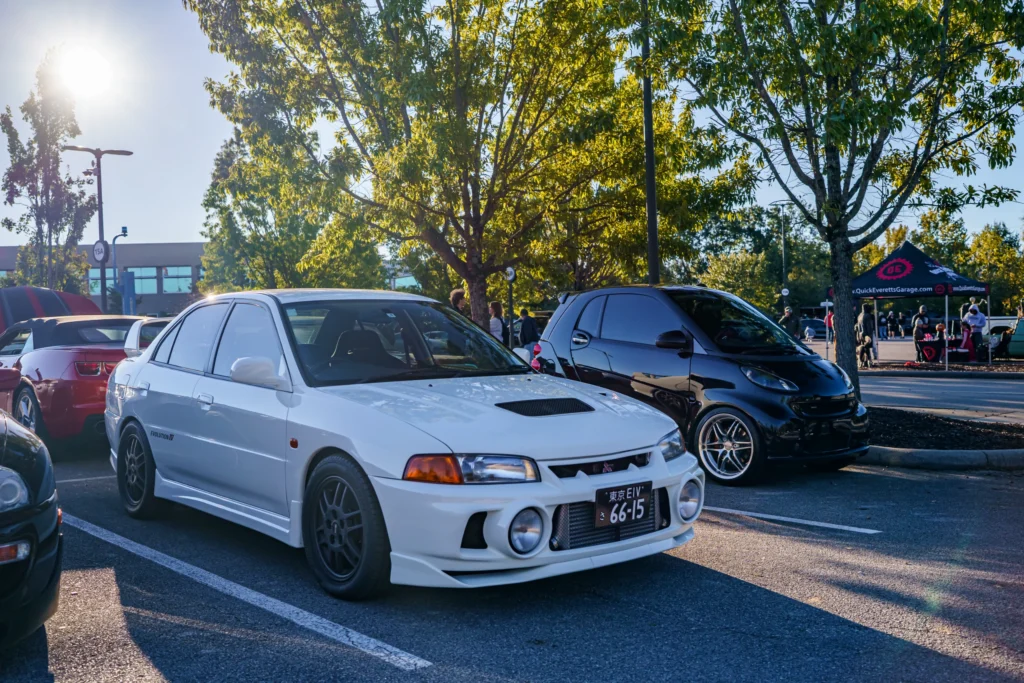 White Mitsubishi Lancer Evolution IV parked in a lot next to a black Smart car and a red convertible with trees and people
