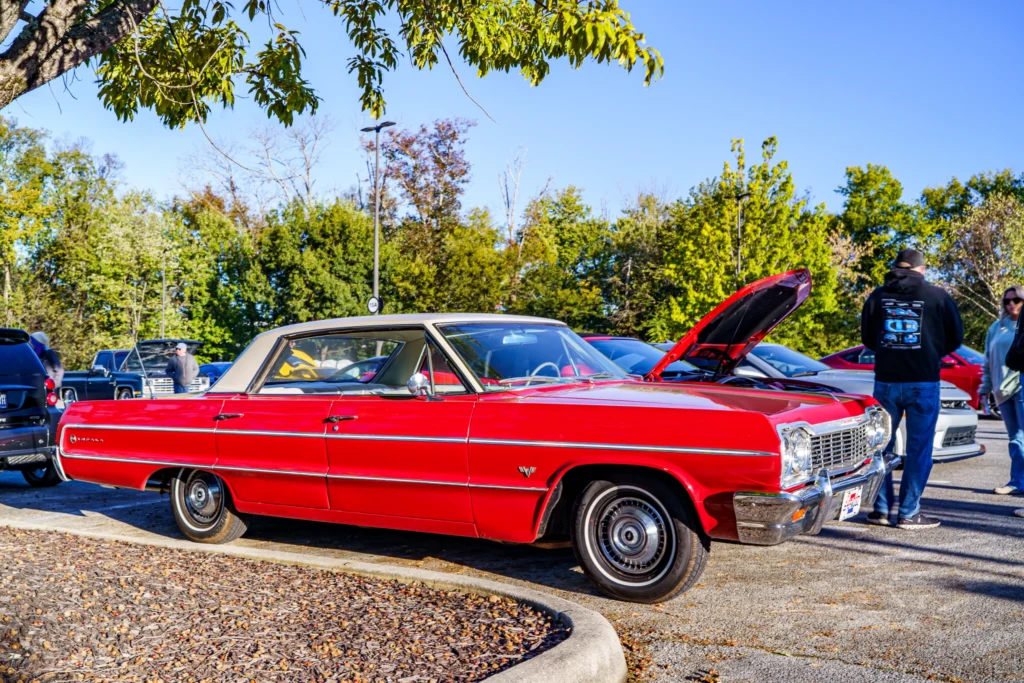 Red Chevrolet Impala with white hardtop parked at a car meet on a clear day with people nearby