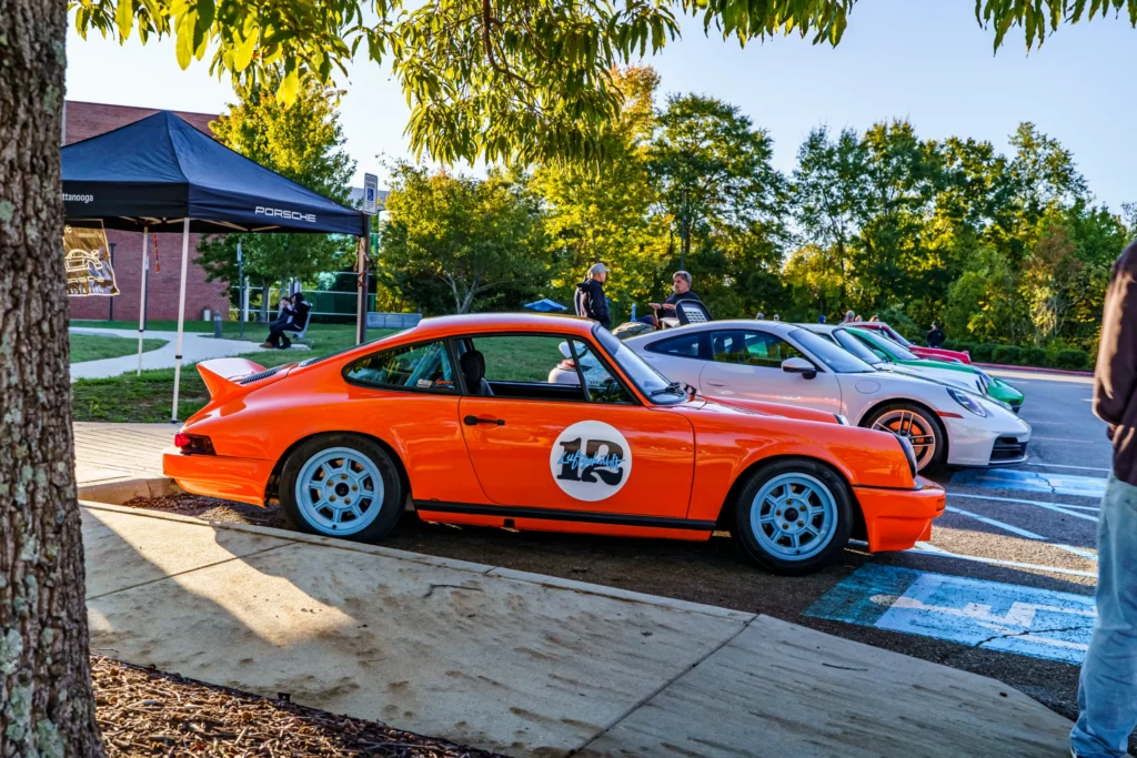 Orange Porsche 911 with white wheels parked beside a line of vehicles at a car event on a sunny day