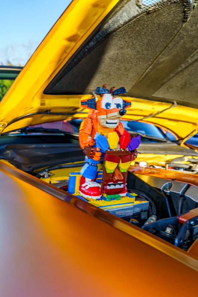 Colorful LEGO Crash Bandicoot of an animated cartoon raccoon on the engine bay of a yellow car with the hood open.