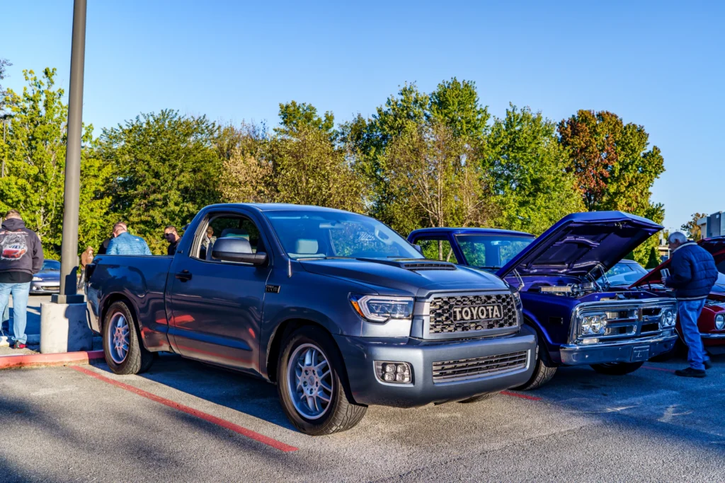 A gray Toyota Tundra is parked beside a blue pickup with its hood raised at an outdoor car show.