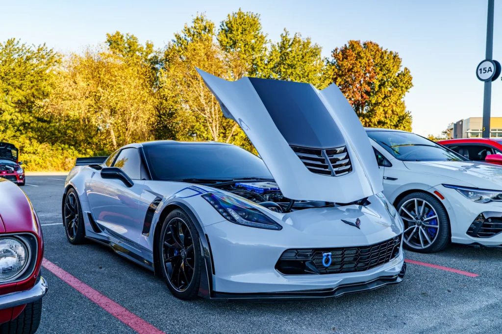 White Chevrolet Corvette with open hood in a parking lot at a car meet with trees in the background