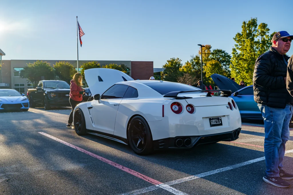 White Nissan GT-R parked at a car event with other vehicles and people around in a sunny outdoor lot