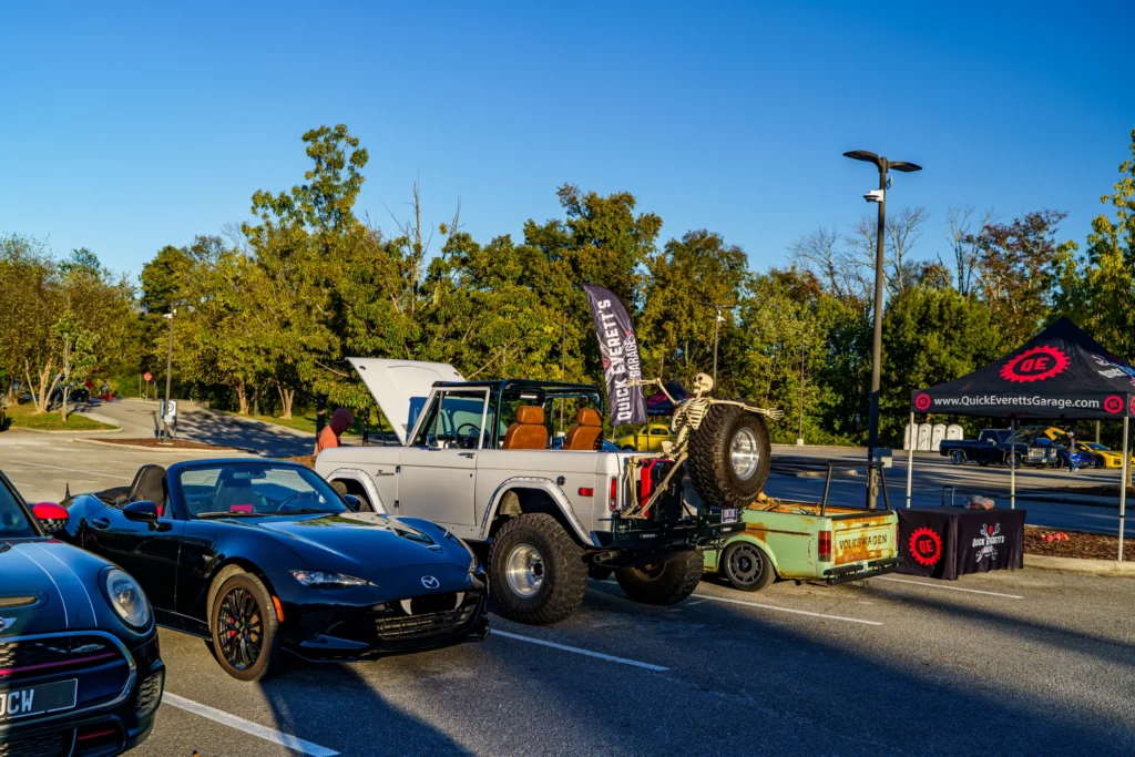 Black Mazda MX-5 Miata convertible with a white Ford Bronco SUV and a green Volkswagen pickup in a parking lot at a car event