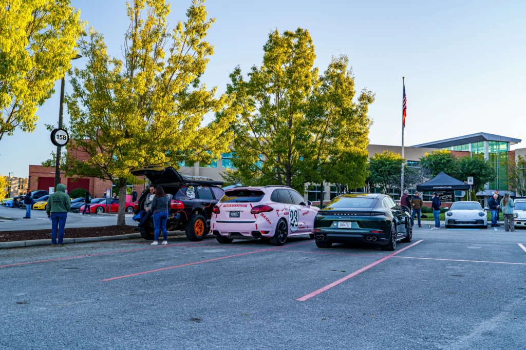 Attendees gather around a green Porsche Panamera, pink and black Cayennes, and a white 911 in a tree-lined parking lot.