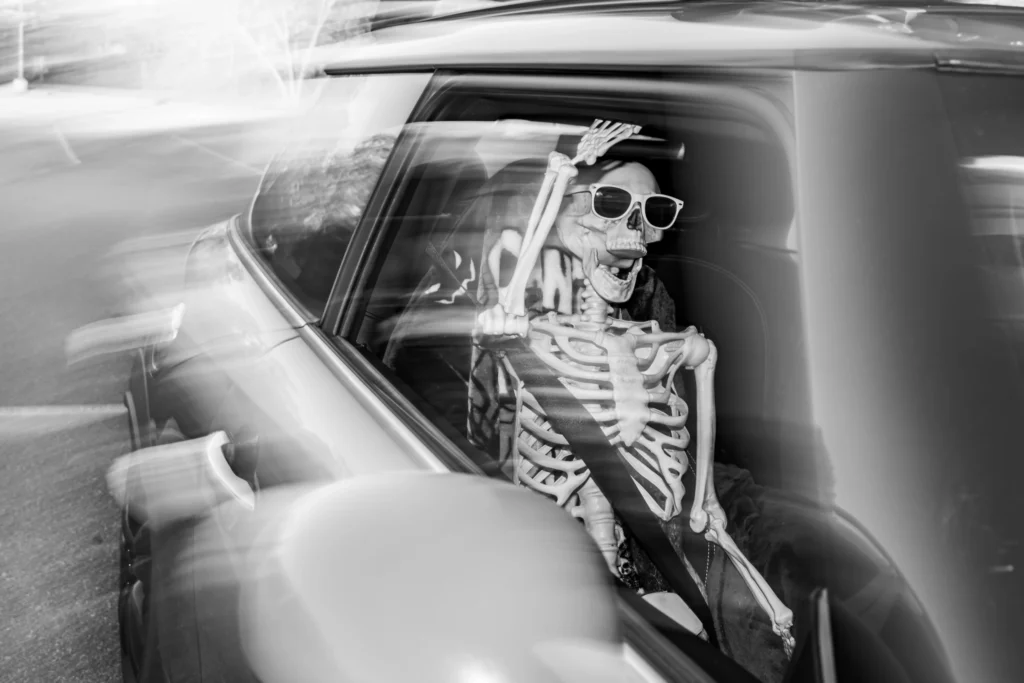 Skeleton figure wearing sunglasses seated in the driver's seat of a car with motion blur outside the window