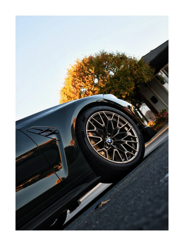 A black BMW's front wheel and fender are shown close up beside a building and a tree with autumn leaves.